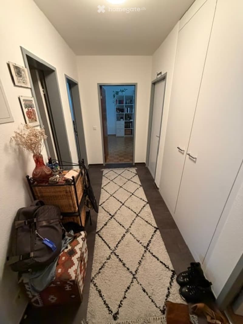 A narrow hallway with white walls, a patterned rug, and several doors leading to other rooms. There are some items like a backpack and shoes placed on the floor.