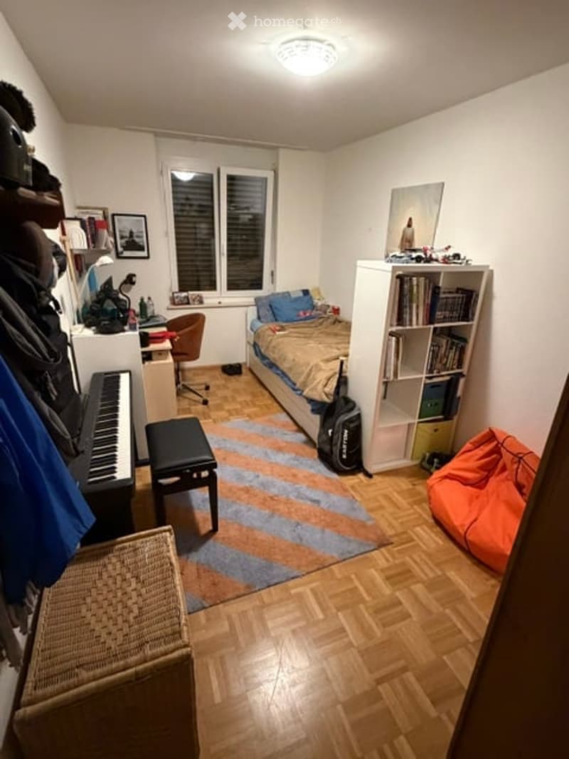 Cozy bedroom with wooden flooring, a bed with pillows and blankets, a bookshelf filled with books, a small black stool, a chair, a piano, a wicker basket, and a beanie bag in orange.