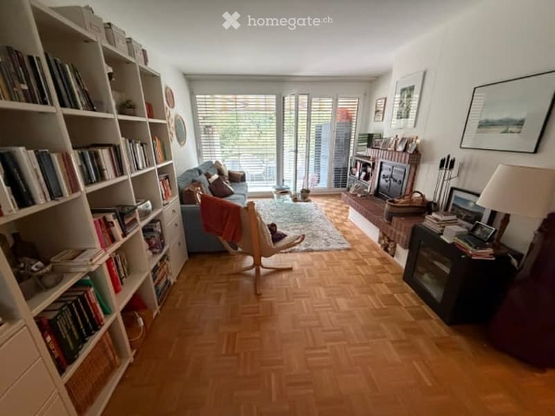 Living room with wooden parquet flooring, large windows, balcony access, sofa, bookshelf, chair, table lamp, fireplace, and picture frames.