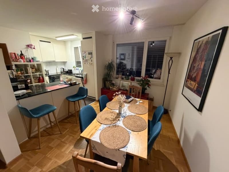 Open-plan living and dining area with modern furniture, including a dining table with six chairs, a kitchen counter with stools, and shelves with various items. The space features wooden flooring, white walls, and large windows with blinds.