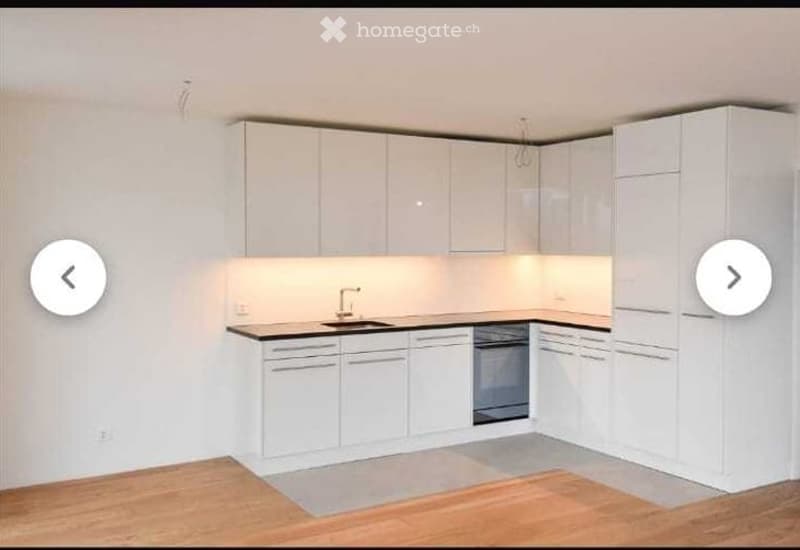 Modern, minimalist kitchen with white cabinets, a black countertop, and integrated appliances including a sink, faucet, and oven. The space features light wood flooring and subtle under-cabinet lighting.