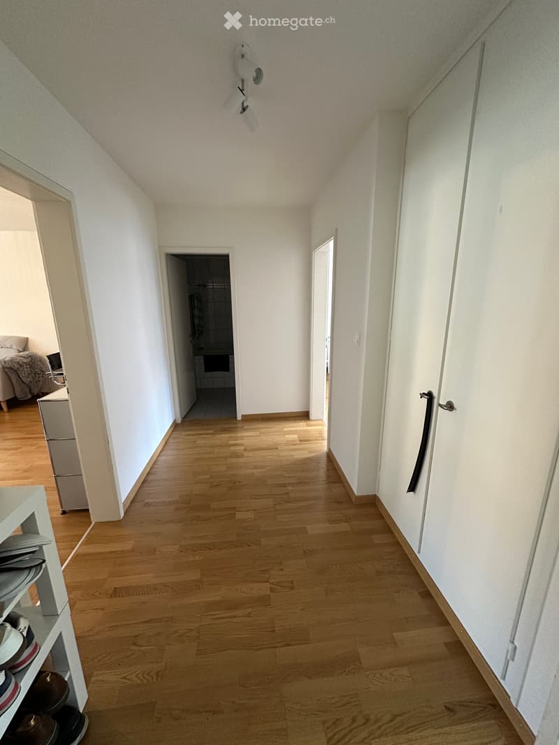 A narrow hallway with wooden flooring, white walls, and a ceiling light fixture. The hallway leads to a bedroom and a bathroom, with a shoe rack on the left side.