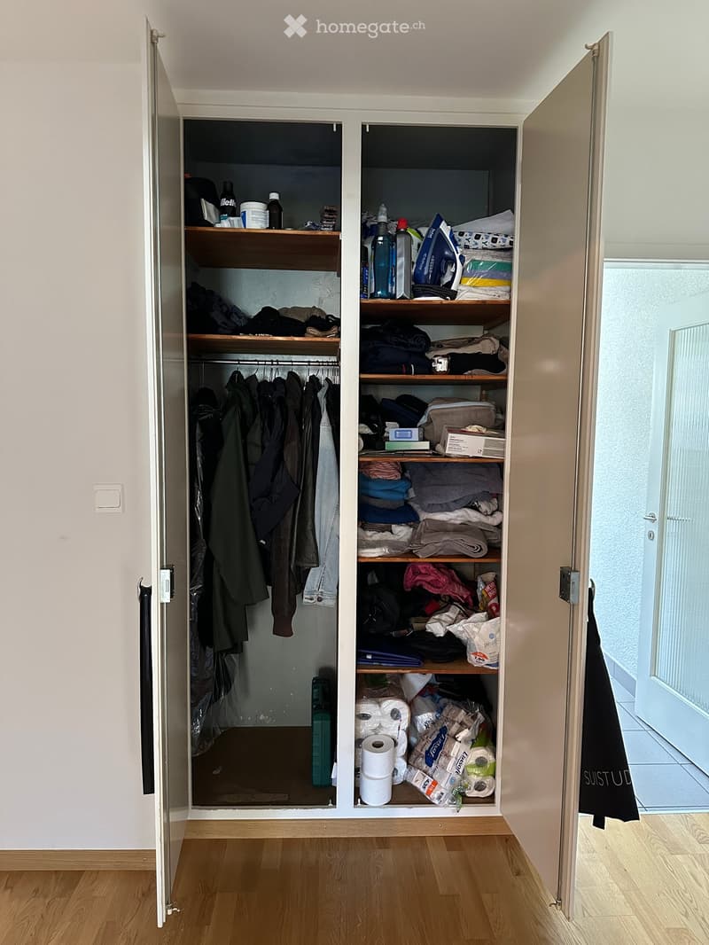 Open closet with wooden shelves and hanging space, containing folded clothes, bottles, and other stored items. The room has light wooden flooring and white walls.