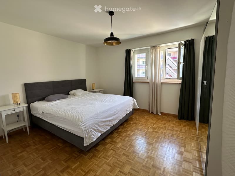 Modern bedroom with a large bed, nightstand, and a hanging pendant light. The room features wooden flooring and large windows with curtains.