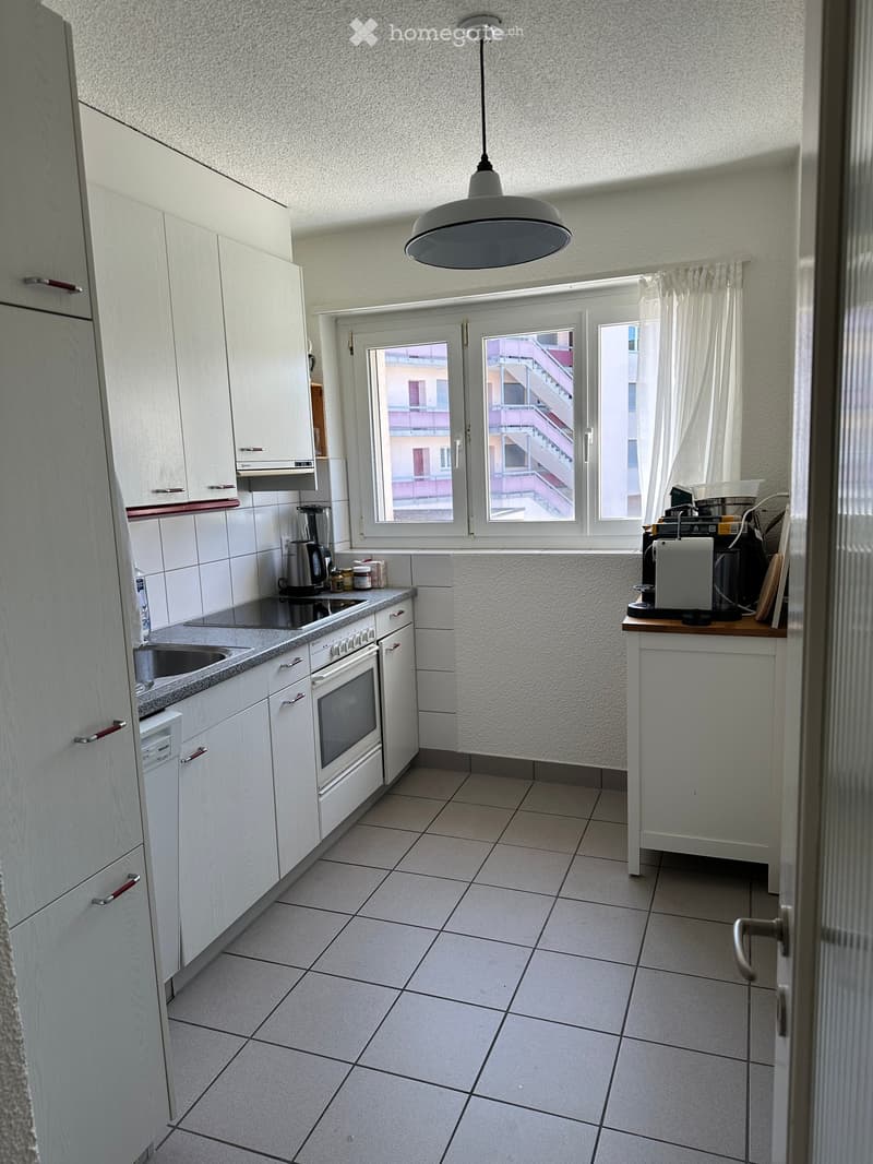 Modern kitchen with white cabinets, tiled floor, and stainless steel appliances including an oven, sink, and coffee maker. A window offers a view of a staircase in the building.