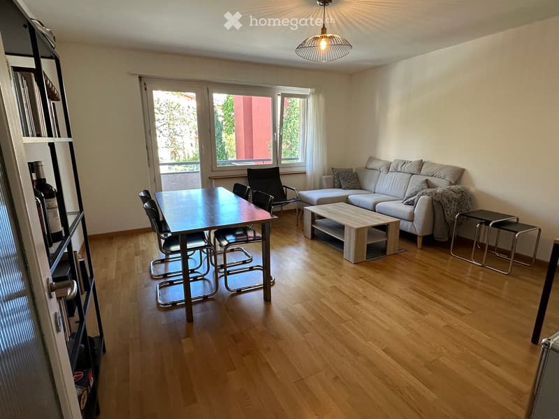 Modern living room with light wooden flooring, large windows, grey sofa, coffee table, dining table with chairs, and a shelf with bottles.