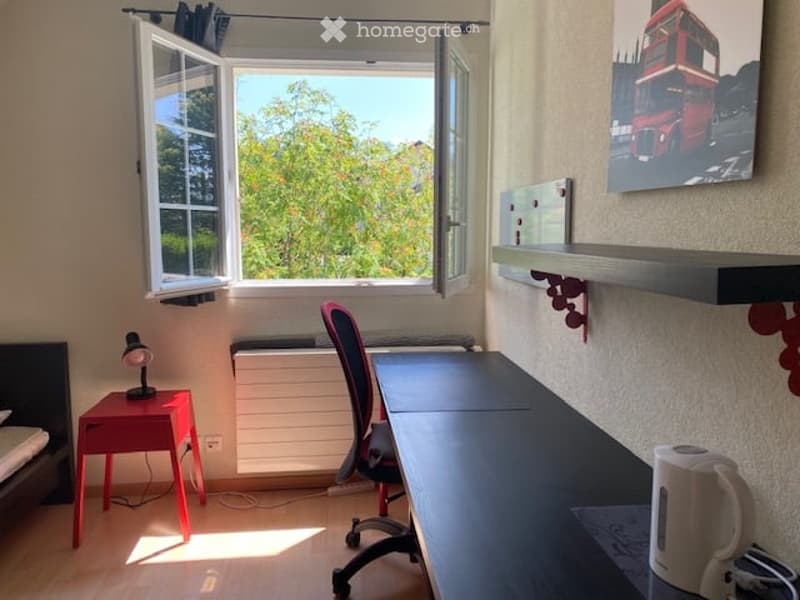 Cozy, well-lit room with a bed, desk, chair, and a small red side table with a lamp. Large windows offer a view of greenery outside.