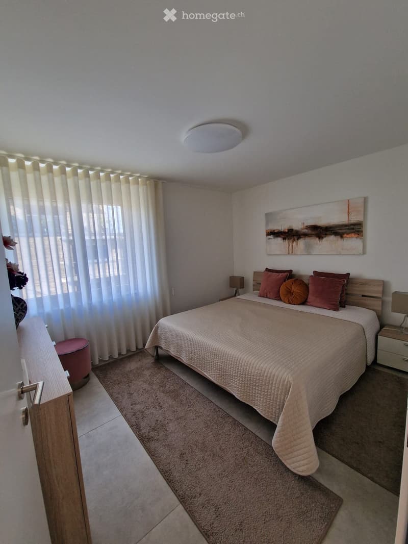 Modern bedroom with a large bed, nightstands, lamps, a round stool, and a painting on the wall. Light-colored curtains cover the windows, and a soft area rug is placed on the floor.