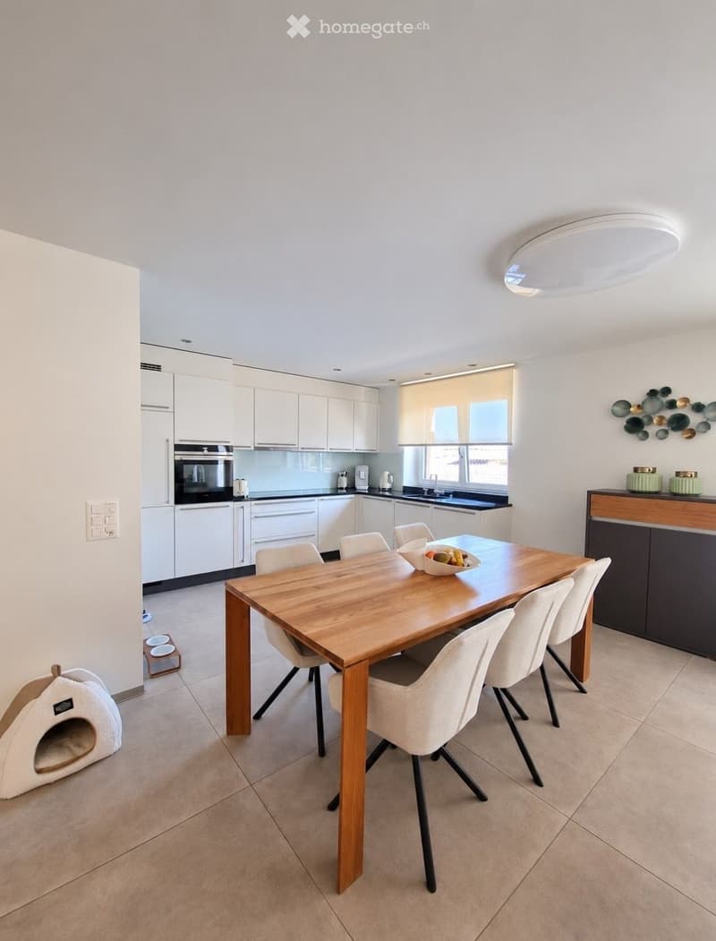 Modern dining area with a wooden dining table, white chairs, a bowl of fruit, and a cat bed. The adjacent kitchen features white cabinets, an oven, and a sink.