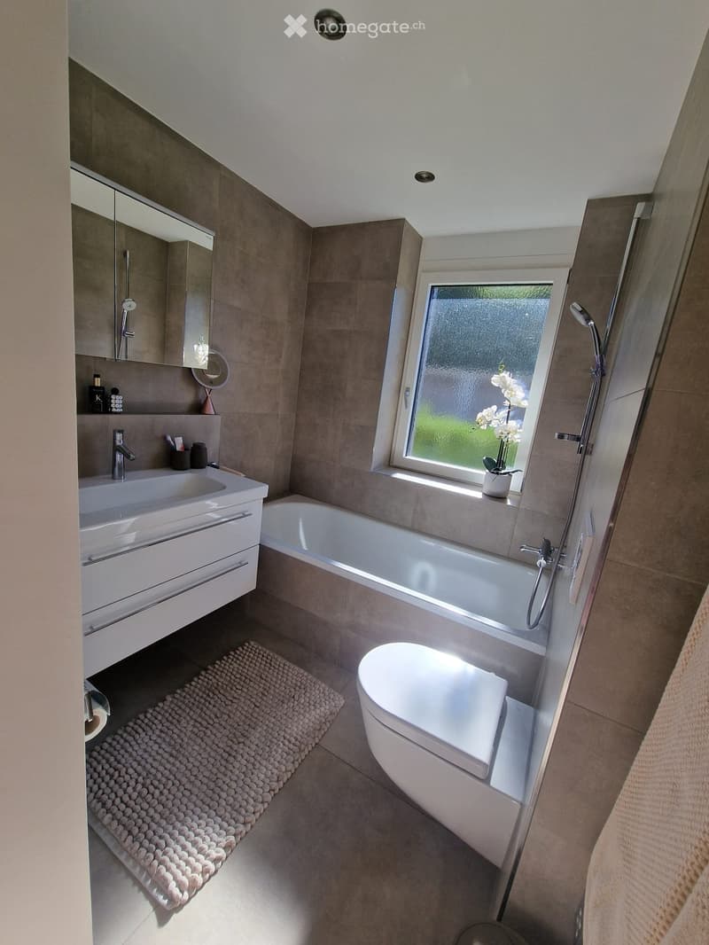 Modern bathroom with a white sink, bathtub, toilet, mirror, and a window with a view of greenery. Includes a showerhead and a small flower vase on the windowsill.