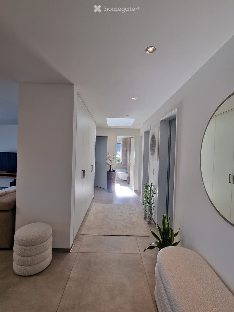 Modern hallway with white walls, tiled floor, recessed lighting, and a large round mirror on the right wall. Features a small rug, potted plants, and a view into a bathroom at the end.