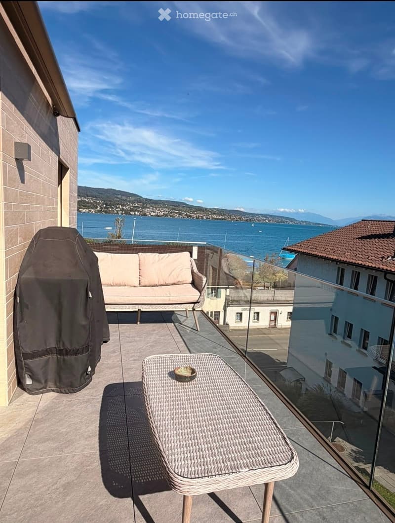 Balcony with a view of a lake and mountains, featuring a sofa and a wicker table.