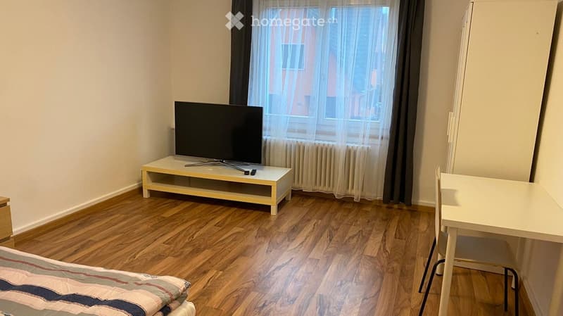 A furnished room with a bed, a small table and chair, a television on a stand, and a window with curtains.