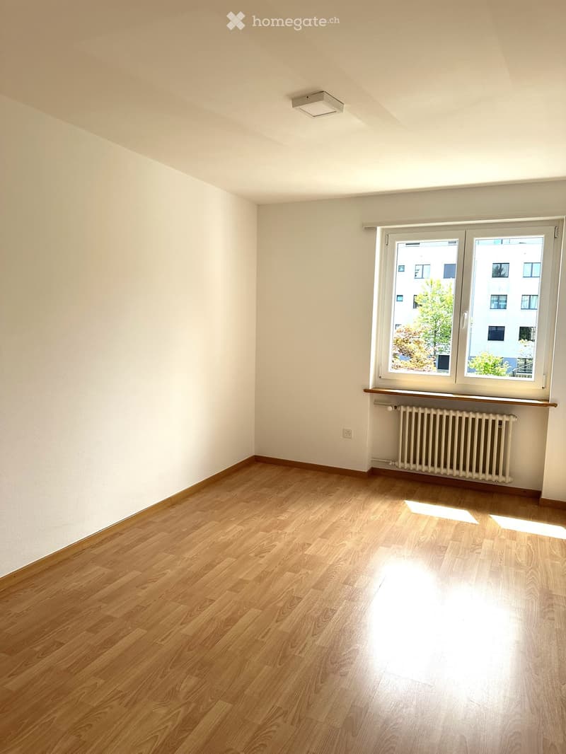 Empty room with light wooden flooring, white walls, and a large window with a radiator below it.