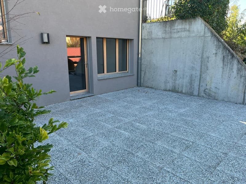 Gravel patio area next to a modern house with large windows and a sliding glass door. A small green plant is visible in the foreground.