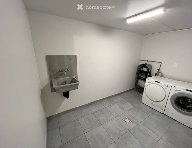 A small bathroom with a sink, washing machine, and dryer. The walls are white, and the floor is tiled.