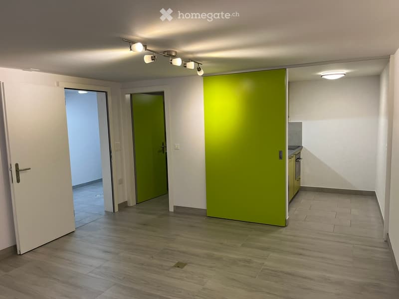 Empty hallway with wooden flooring, white walls, and a green accent wall. Features track lighting, a partially open door leading to another room, and a small kitchen area visible at the end.
