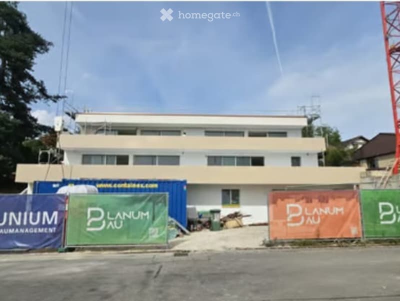 A two-story building with a flat roof and multiple windows, surrounded by construction banners and a partially visible construction site.