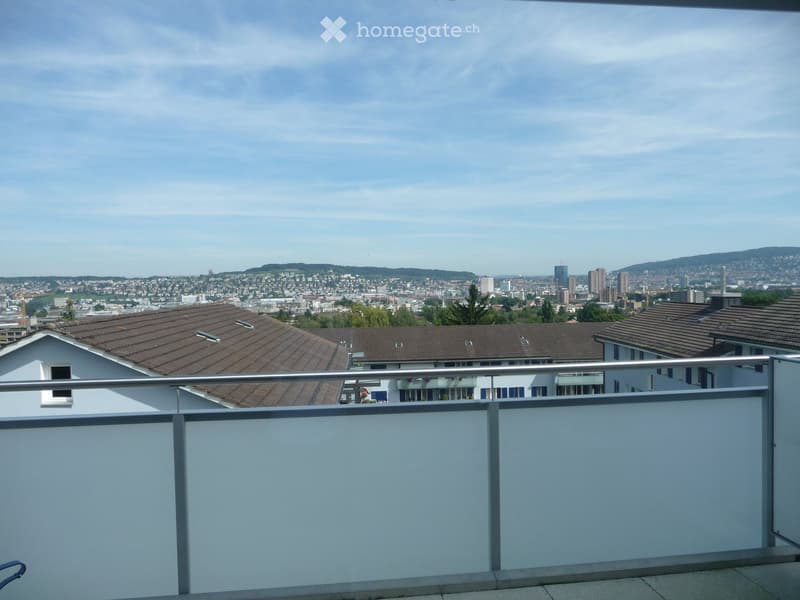 Balcony with metal railing overlooking a cityscape with residential buildings, trees, and distant hills under a partly cloudy sky.