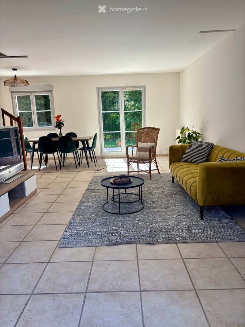 Bright living room with tiled floor, large windows, and a balcony door leading outside. Features a green velvet sofa, a black coffee table, a wicker armchair, a dining table with chairs, and a television on a wooden stand.
