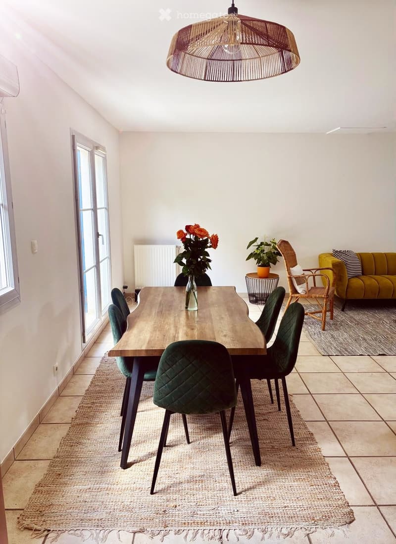 Dining area with a wooden dining table, green upholstered chairs, a yellow sofa, a wicker pendant light, and potted plants.