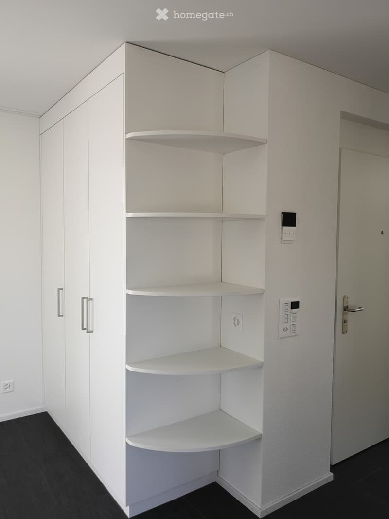 Corner of an unfurnished room with white built-in shelving unit, white cabinets, and a door.
