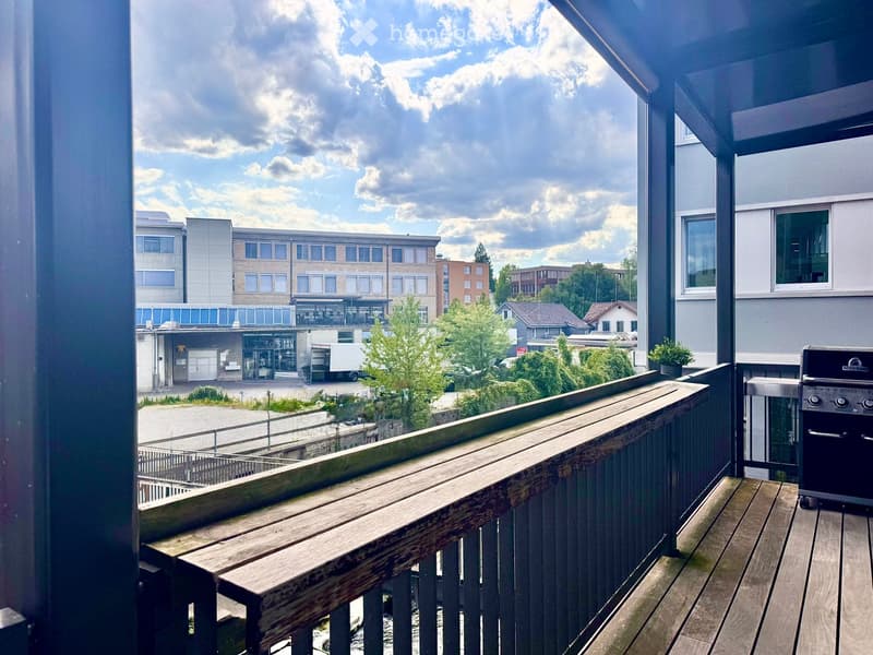 Balcony with wooden decking, railing, and a grill, overlooking a modern building and greenery.