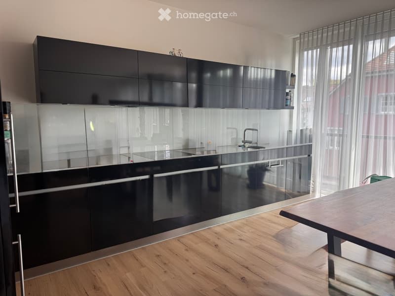 Modern kitchen with black cabinets, glass backsplash, stainless steel sink, and wooden floor.