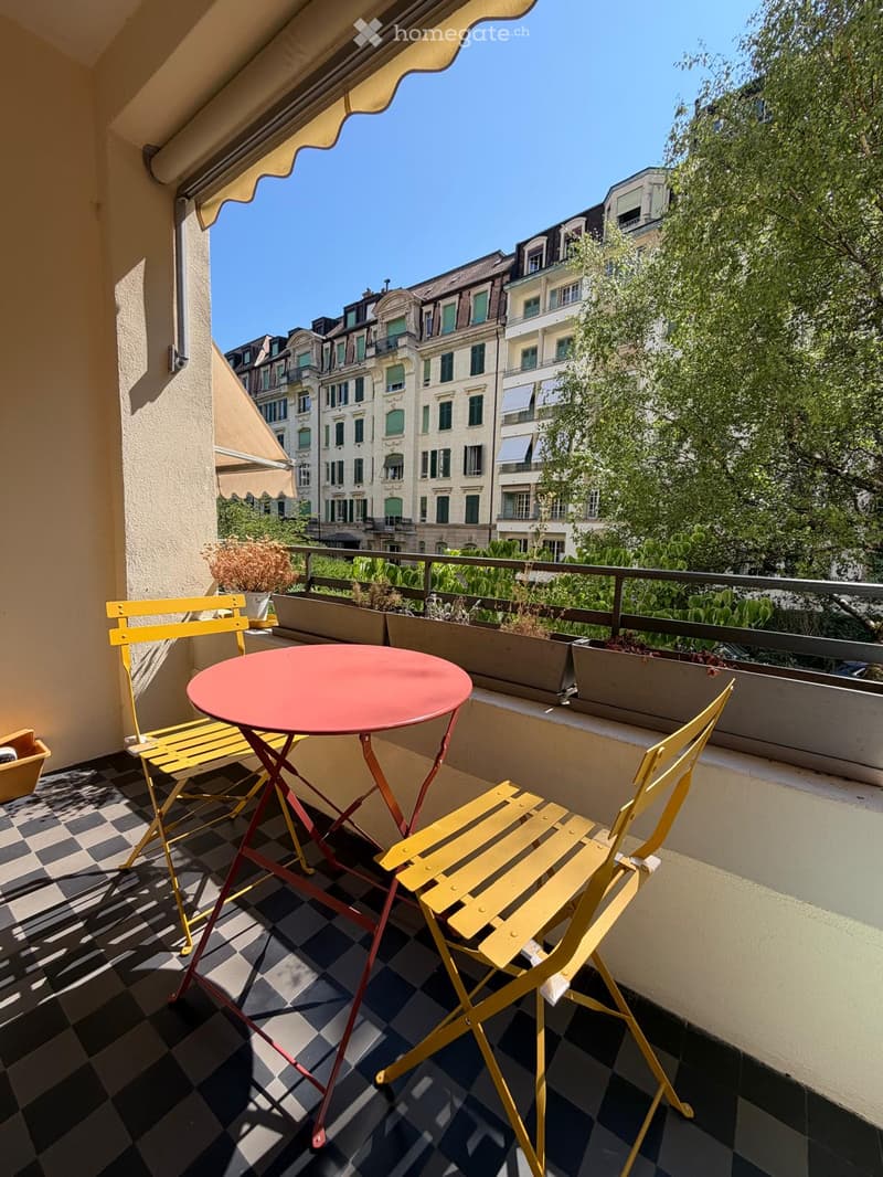 Balcony with a red round table and two yellow chairs, checkered floor, potted plants, and a view of city buildings.