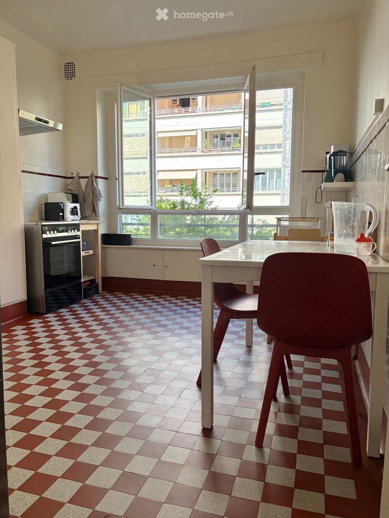 Bright kitchen with checkered red and white tiles, white walls, and large windows overlooking a balcony. Includes a table with chairs, an oven, coffee maker, and other appliances.