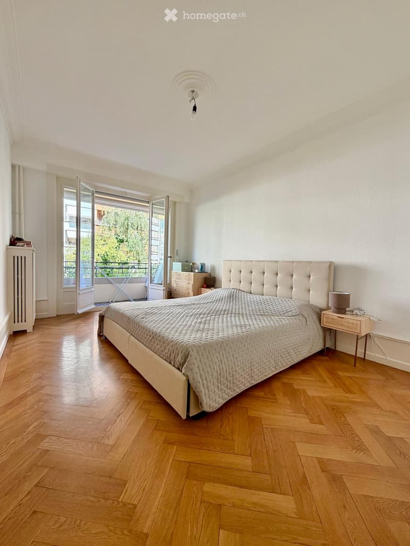 Spacious bedroom with wooden flooring, large bed with tufted headboard, nightstand with lamp, radiator, and open balcony door leading to a terrace.