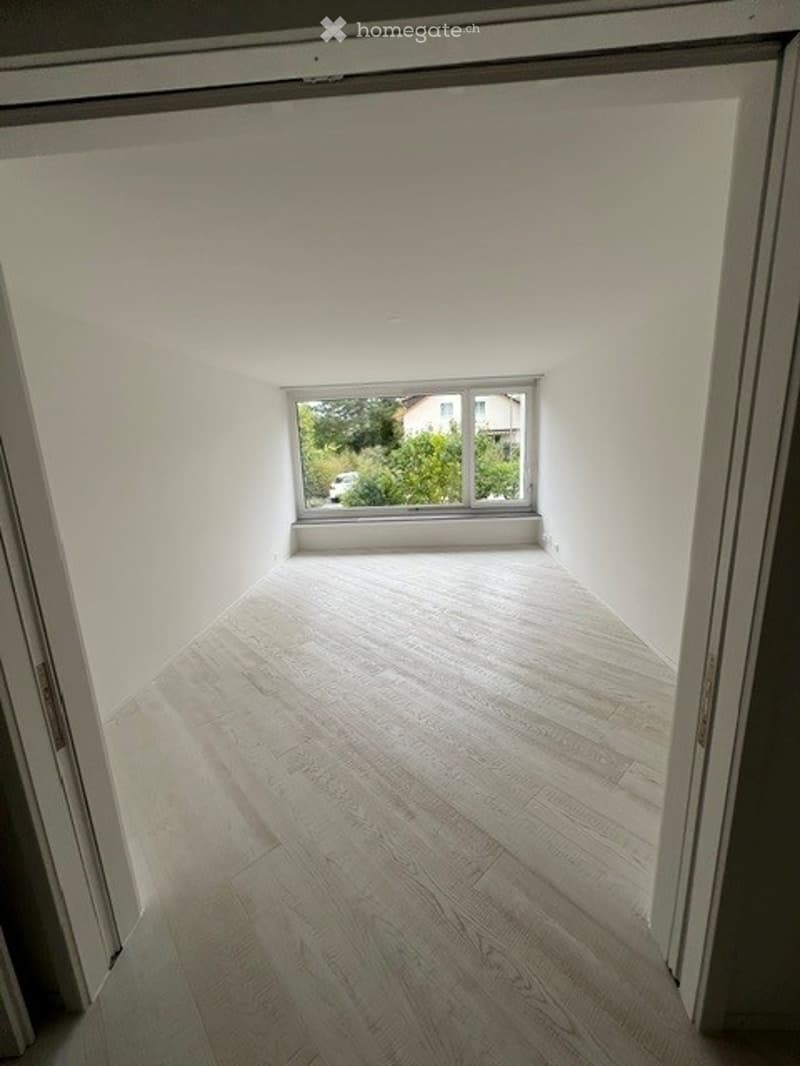 Empty room with light wooden flooring and white walls, featuring a large window with a view of greenery outside.
