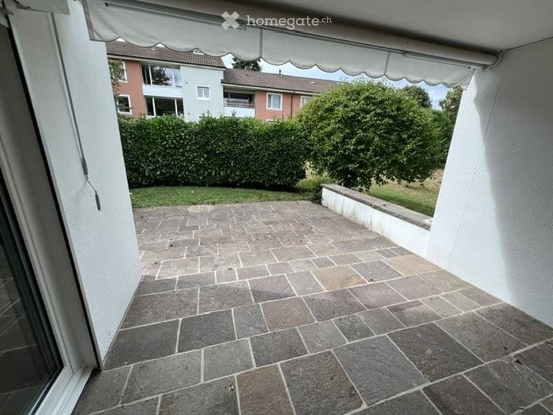 Patio with stone tiles, covered by a retractable awning, surrounded by a low wall, with a neatly trimmed hedge and trees in the background.
