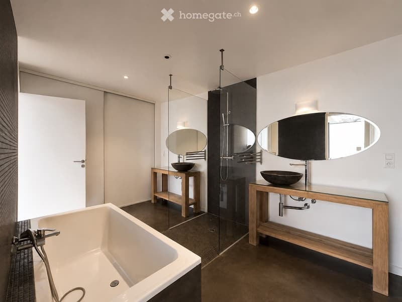 Modern bathroom with a freestanding bathtub, two wooden sink vanities, oval mirrors, and a glass-enclosed shower area.