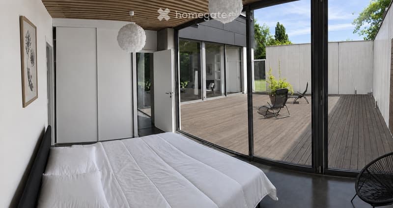 Modern bedroom with a large bed, white bedding, and a sliding glass door leading to a wooden deck with outdoor seating.