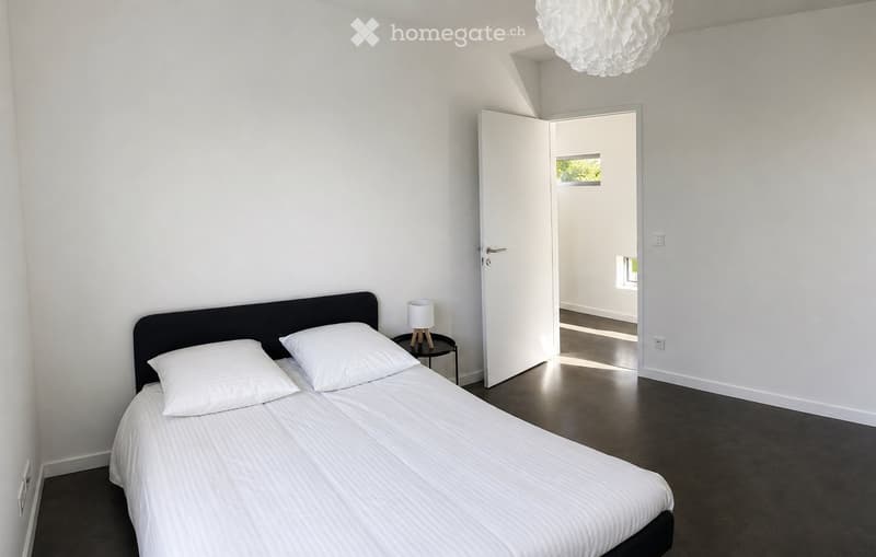 Modern bedroom with a double bed, white bedding, a nightstand with a lamp, and an open door leading to another room.
