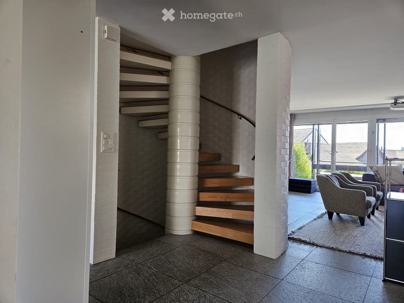 Modern living room with a curved staircase, white brick walls, grey sofa, chairs, ottoman, and a large window with a view.