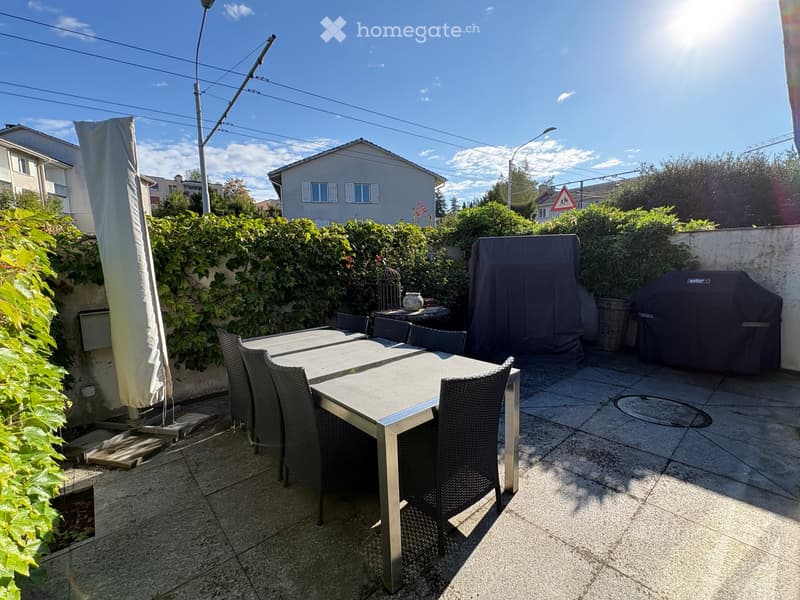 Outdoor patio with a dining table, chairs, umbrella, and covered grill, surrounded by greenery and a clear sky.