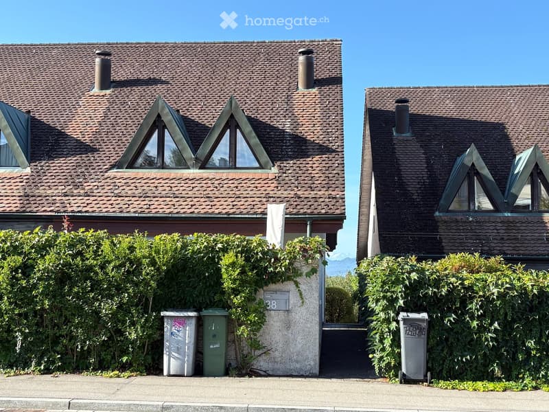 Two-story house with brown tiled roof, chimney, and triangular dormer windows. Green bushes surround the house, and there are trash bins in front.