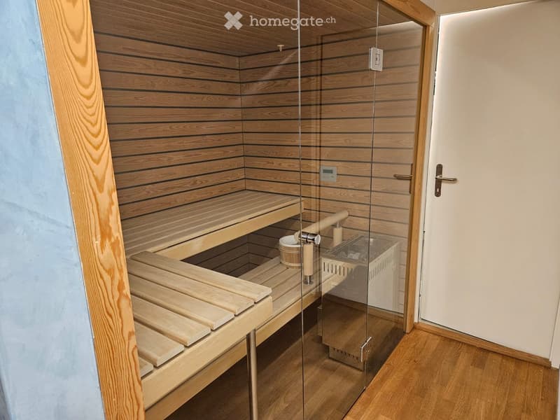 Wooden sauna with multiple levels of benches, a wooden bucket, and a glass-enclosed electric heater. The sauna features light wooden paneling and a control panel on the wall.