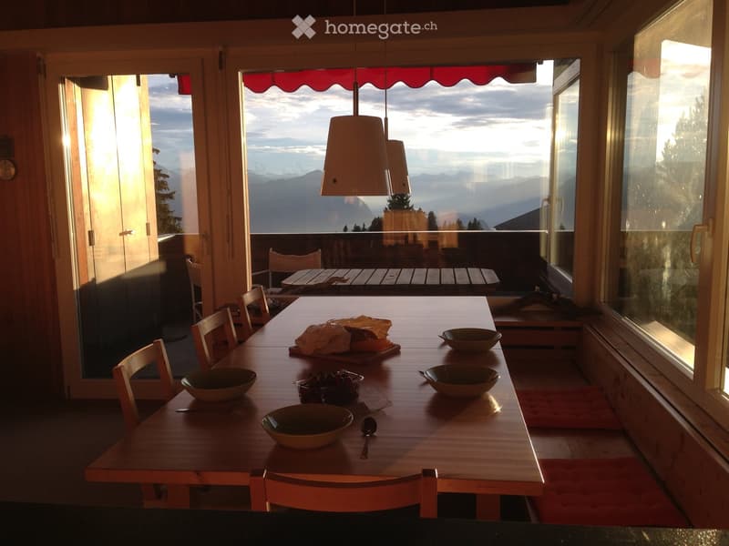 Bright kitchen with large windows offering a scenic mountain view, featuring a wooden dining table set with bowls and a spoon, surrounded by chairs.
