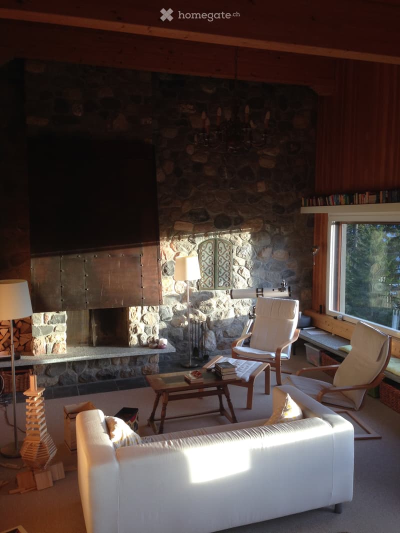 Cozy living room with a stone fireplace, white leather sofa, coffee table, and wooden chairs. Large windows provide natural light.