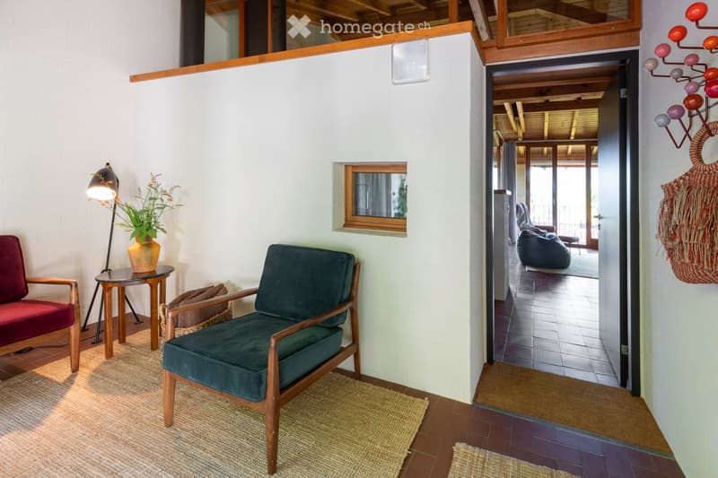 A cozy living room with a red sofa, green armchair, small round table with a lamp and vase, and a view into another room with a bean bag chair.