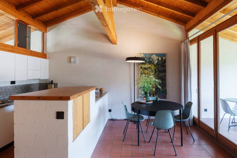 Open-plan kitchen and dining area with wooden bar counter, white cabinets, modern chairs, round dining table, floor lamp, and artwork on the wall.