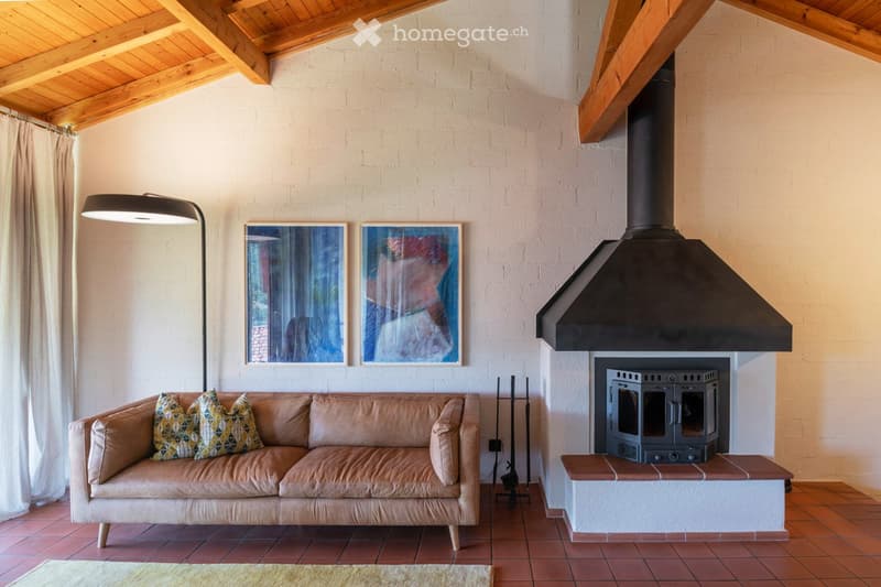 Living room with exposed wooden beams, a tan leather sofa with patterned pillows, a floor lamp, and a fireplace with artwork above it.