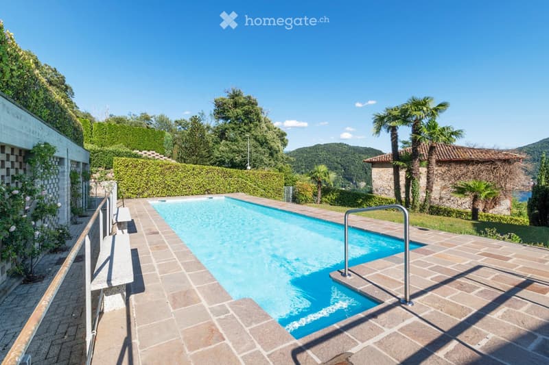 A rectangular swimming pool surrounded by a tiled patio, with a metal handrail on one side, lush greenery, and a view of a hillside and distant water.