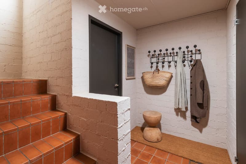 A modern hallway with exposed brick walls, terracotta tiled steps, a coat rack with a woven basket, a small wooden stool, and a framed picture on the wall.