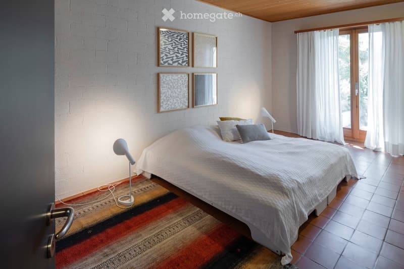 Modern bedroom with a white brick wall, a large bed with pillows, a colorful rug, and two table lamps on either side of the bed. There are two mirrors and picture frames on the wall above the bed.