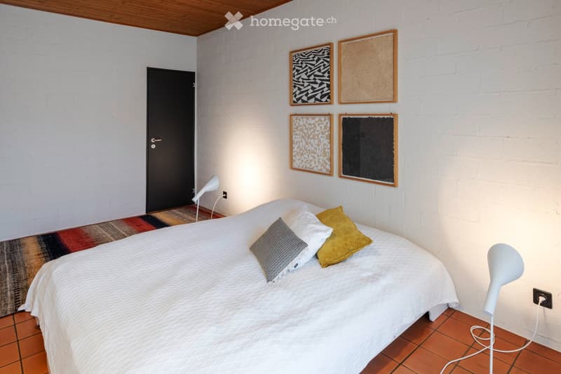 Modern bedroom with white brick walls, a wooden ceiling, a bed with pillows, and decorative picture frames on the wall.