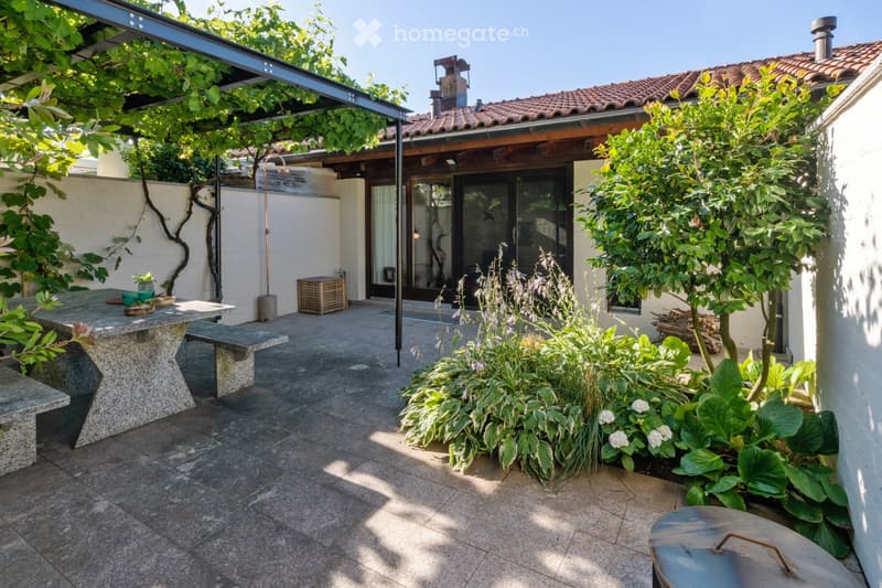 A well-maintained outdoor patio area with a stone table, benches, lush greenery, and a covered section leading to a house entrance.